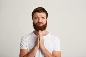 Closeup of peaceful handsome young man hipster with beard wears t shirt feels calm and keeps hands folded in praying position isolated over white background photo