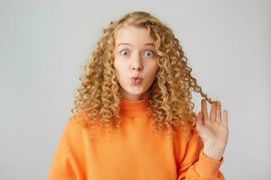 Gorgeous European curly blonde woman pretends to be kissing doing fish face,crazy and comical gesture, stares in camera holding in her hand a strand,isolated on white background photo