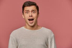 Photo of young scared guy in blank long sleeve, stands over pink background with wide open eyes and screaming, looks crazy and unhappy.