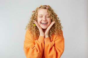 Portrait of a cheerful beautiful girl wearing orange sweater keeps palms near face celebrating with eyes closed in pleasure isolated over white background photo