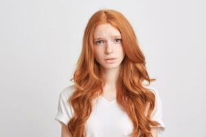 Portrait of upset depressed young woman with red wavy long hair and freckles wears t shirt feels worried and unhappy isolated over white background photo