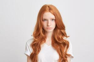 Closeup of beautiful redhead young woman with wavy long hair and freckles wears t shirt feels sad and looks directly in camera isolated over white background photo