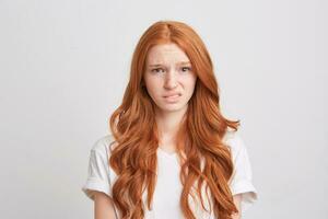 Closeup of sad upset young woman with wavy long hair and freckles wears t shirt feels depressed and looks displeased isolated over white background photo