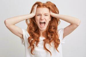 Portrait of desperate hysterical young woman with red wavy long hair, freckles and opened mouth wears t shirt feels angry and shouting isolated over white background photo