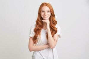 Portrait of smiling charming young woman with red wavy long hair and freckles wears t shirt feels happy and keeps hands folded isolated over white background photo