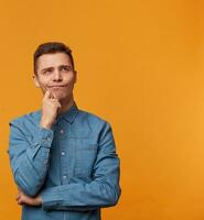 Vertical photo of a pensive, dreamy, attractive man in a trendy denim shirt looking into the distance, holding his hand near his chin, isolated against a yellow background.