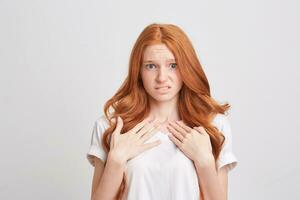 Portrait of sad displeased young woman with red wavy long hair and freckles wears t shirt feels confused and points at herself wih both hands isolated over white background photo