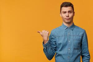 A contented pompous, arrogant guy isolated over yellow background casually draws your attention to what is on the left, shows with thumb finger on the empty space to copy. photo