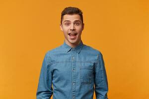 Pleasantly surprised, delighted by the good news, a young guy who happened to meet an old acquaintance, a young guy dressed in a fashionable denim shirt, isolated against a yellow background. photo