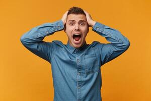 A man in a panic grabbed his head, loudly screaming, a defeat collapse fail had happened, wearing a denim shirt isolated on a yellow background. photo