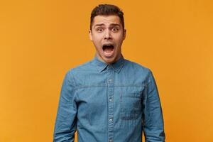 Portrait of a guy who screams in a panic, screaming with fear, makes a loud cry as he is amazed and very scared, wearing a fashionable denim shirt, isolated on a yellow background photo