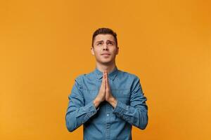 Young guy looks up with hope, palms folded in a prayer gesture, asking for help from higher powers, protection, blessing, wearing a denim shirt, isolated on a yellow background photo