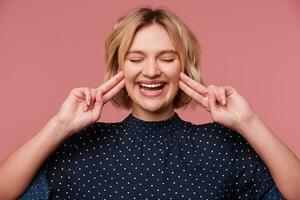 Close up of smiling laughing young attractive blonde woman dressed in blouse with polka dots, closed eyes. keeps two fingers near the ears, showing gesture of deafness, ignores someone, over pink wall photo