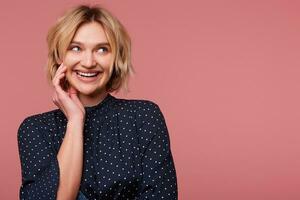 Lively attractive beautiful blonde girl with short haircut looking at right upper corner barely touches her cheek dressed in blouse with polka dots, coquets, flirting, isolated over pink background photo