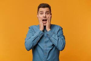 Shocked guy in a panic clutched at his face dressed in a denim shirt, isolated on a yellow background. Astonished omg face expression photo