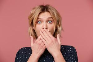 Close up of scared woman having troubles. Frightened blonde in despair covered mouth with both palms,popping eyes keeps silence, secret, dressed blouse with polka dots, isolated over pink background photo