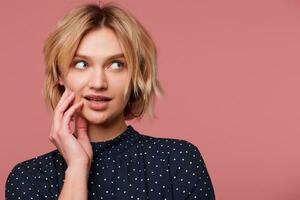 Dreaming attractive beautiful blonde girl with short haircut looking at right upper corner barely touches her cheek dressed in blouse with polka dots, coquets, flirting, isolated over pink background photo