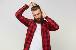 Handsome bearded young man in shirt doing modern hairstyle, grooming his hair with comb isolated over white background photo