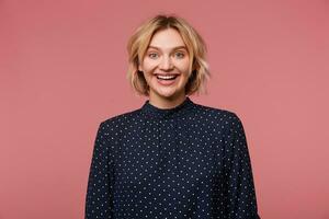 Portrait of young beautiful attractive blonde woman dressed in blouse with polka dots, has exited face expression, showing positive, smiling, happy, isolated over pink background photo