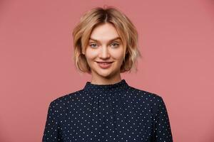 Blue-eyed young pretty blonde woman has surprised facial expression, dressed in blouse with polka dots, showing amazement, isolated over pink background photo