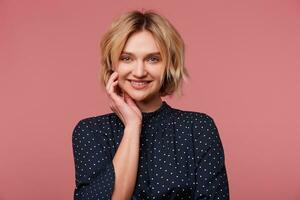 Portrait of attractive flirtatious young beautiful blonde with short haircut keeping hand near the face, dressed in blouse with polka dots,coquets, flirting, smiling pleasantly,isolated over pink wall photo