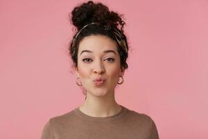 Portrait of charming girl with dark curly hair bun. Wearing headband, earrings and brown sweater. Has make up. Purses lips in a kiss. Watching at the camera isolated over pastel pink background photo