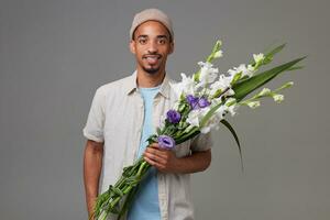 retrato de joven alegre atractivo chico en gris sombrero, sostiene un ramo de flores en su manos, mira a el cámara con contento expresión y sonriente, soportes terminado gris fondo foto