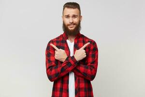 Young bearded man in checkered shirt with fashion hairstyle with arms crossed shows the index fingers right and left, doubts does not know what to choose, hard to make a decision, on white background photo