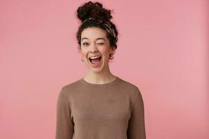 Joyful girl, happy looking woman with dark curly hair bun. Wearing headband, earrings and brown sweater. Has make up. Emotion concept. Winks at the camera isolated over pastel pink background photo