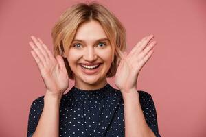 Close up of excited young beautiful attractive blonde woman dressed in blouse with polka dots, keeps palms near the face has exited face expression, showing positive, smiling, happy, over pink wall photo