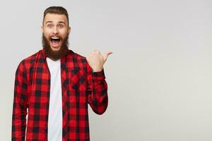 Portrait of cheerful attractive joyful bearded man in checkered shirt with mustache fashion hairstyle, opened mouth because of amazement pointing on copyspace behind his back isolated on white wall photo