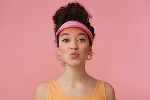 Lovely girl, flirty woman with dark curly hair bun. Wearing pink visor, earrings and orange tank top. Has make up. Purses lips in a kiss. Watching at the camera isolated over pastel pink background photo