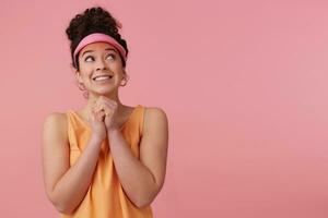 Asking, cute woman with dark curly hair bun. Wearing pink visor, earrings, orange tank top. Has make up. Keeps palms together. Watching to the right at copy space, isolated over pastel pink background photo