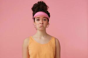 Portrait of sad girl with dark curly hair bun. Wearing pink visor, earrings and orange tank top. Has make up. Emotion concept. Pouts a lip. Watching at the camera isolated over pastel pink background photo