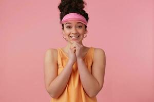 Portrait of pleading girl with dark curly hair bun. Wearing pink visor, earrings and orange tank top. Has make up. Keeps palms together. Watching at the camera isolated over pastel pink background photo