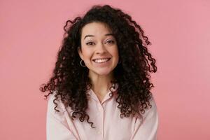 Portrait of attractive, adult girl with long dark curly hair. Wearing earrings and pastel pink shirt. Has make up. Broadly smiling. Watching at the camera isolated over pastel pink background photo