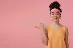 Beautiful girl with dark curly hair bun. Wearing pink visor, earrings and orange tank top. Has make up. Watching at the camera, pointing thumb to the left at copy space, isolated over pink background photo