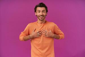 Happy man with dark hair and bristle. Wearing orange sweater with rolled up sleeves. Has bracelets, rings. Pointing at himself with excitement. Watching at the camera isolated over purple background photo