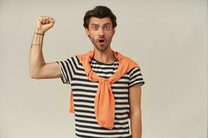 Rebellious guy with dark hair and bristle. Wearing striped t-shirt, sweater tied on shoulders. Raises fist up, protesting. Has bracelets and rings. Watching at the camera isolated over grey background photo