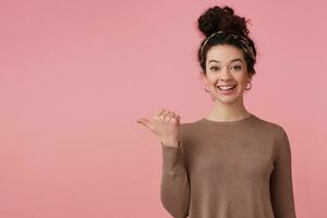 Happy woman with dark curly hair bun. Wearing headband, earrings, brown sweater. Has make up. Watching at the camera and pointing thumb to the left at copy space, isolated over pastel pink background photo