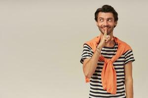 Tricky looking guy with brunette hair and bristle. Wearing striped t-shirt and orange sweater tied on shoulders. Shows silence sign. Watching to the left at copy space, isolated over grey background photo