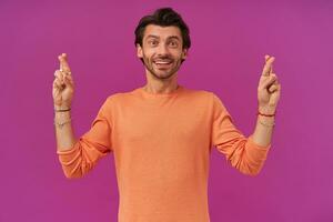 Happy looking guy with brunette hair and bristle. Wearing orange sweater with rolled up sleeves. Has bracelets and rings. Keeps fingers crossed. Watching at the camera isolated over purple background photo