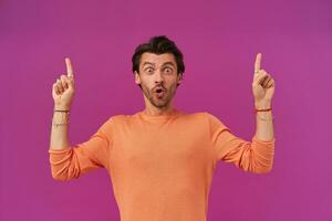 Shocked man with dark hair and bristle. Wearing orange sweater with rolled up sleeves. Has bracelets, rings. Watching at the camera, pointing fingers up at copy space, isolated over purple background photo