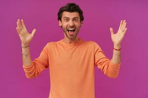 Portrait of excited male with brunette hair and bristle. Wearing orange sweater with rolled up sleeves. Has bracelets and rings. Raises hands. Watching at the camera isolated over purple background photo