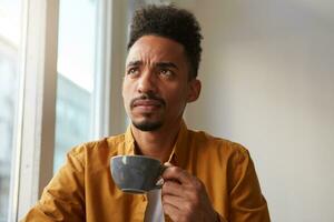 African  man wears in yellow shirt, sitting at a table in a cafe and drinks aromatic coffee, remembers whether the iron turned off before leaving the apartment. Looking thoughtfully into the distance. photo