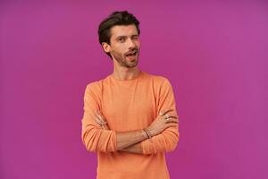 Portrait of flirty male with brunette hair and bristle. Wearing orange sweater with rolled up sleeves. Has bracelets and rings. Keeps arms crossed. Winks at the camera isolated over purple background photo