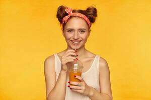 joven dama, bonito jengibre mujer con dos bollos y banda para el cabello mirando contento. vistiendo blanco camisa y participación su sano zalamero. sonriente acecho a el cámara aislado terminado amarillo antecedentes foto