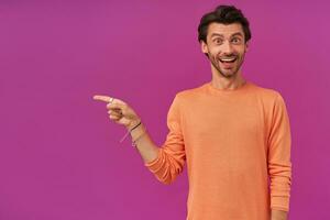 Surprised man with brunette hair and bristle. Wearing orange sweater. Has bracelets and rings. Watching at the camera and pointing finger to the left at copy space, isolated over purple background photo