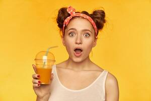 Portrait of attractive, red hair girl with red doted hairband. Wearing white shirt and holding her smoothie, expressing shock. Watching at the camera isolated over yellow background photo