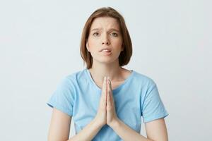 Closeup of upset worried young woman in blue t shirt keeps hands folded in praying position and biting her lip isolated over white background Begging for help photo